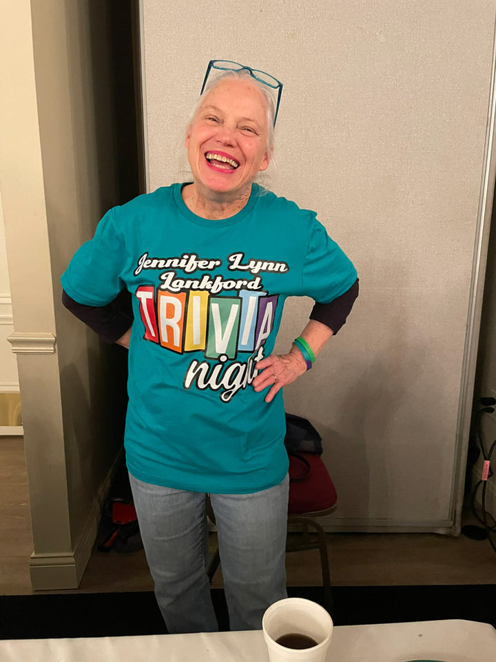 Smiling woman wearing a teal “Jennifer Lynn Lankford Trivia Night” tee in rainbow letters, standing at a community event table.