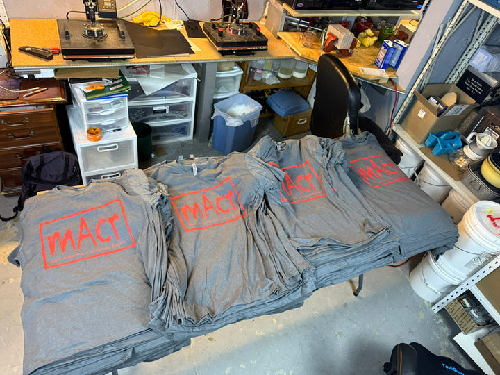 Multiple gray performance tees printed with a red MACT logo and Mid America Collision Center text stacked on a print shop table