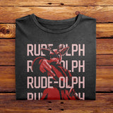 A vintage folded black comfort colors shirt. The retro design features Rudolph the Red Nosed Reindeer in a y2k merch artstyle, posed in a rude manner. There is text that says Rude-olph, a funny festive pun with his name to go with the graphic