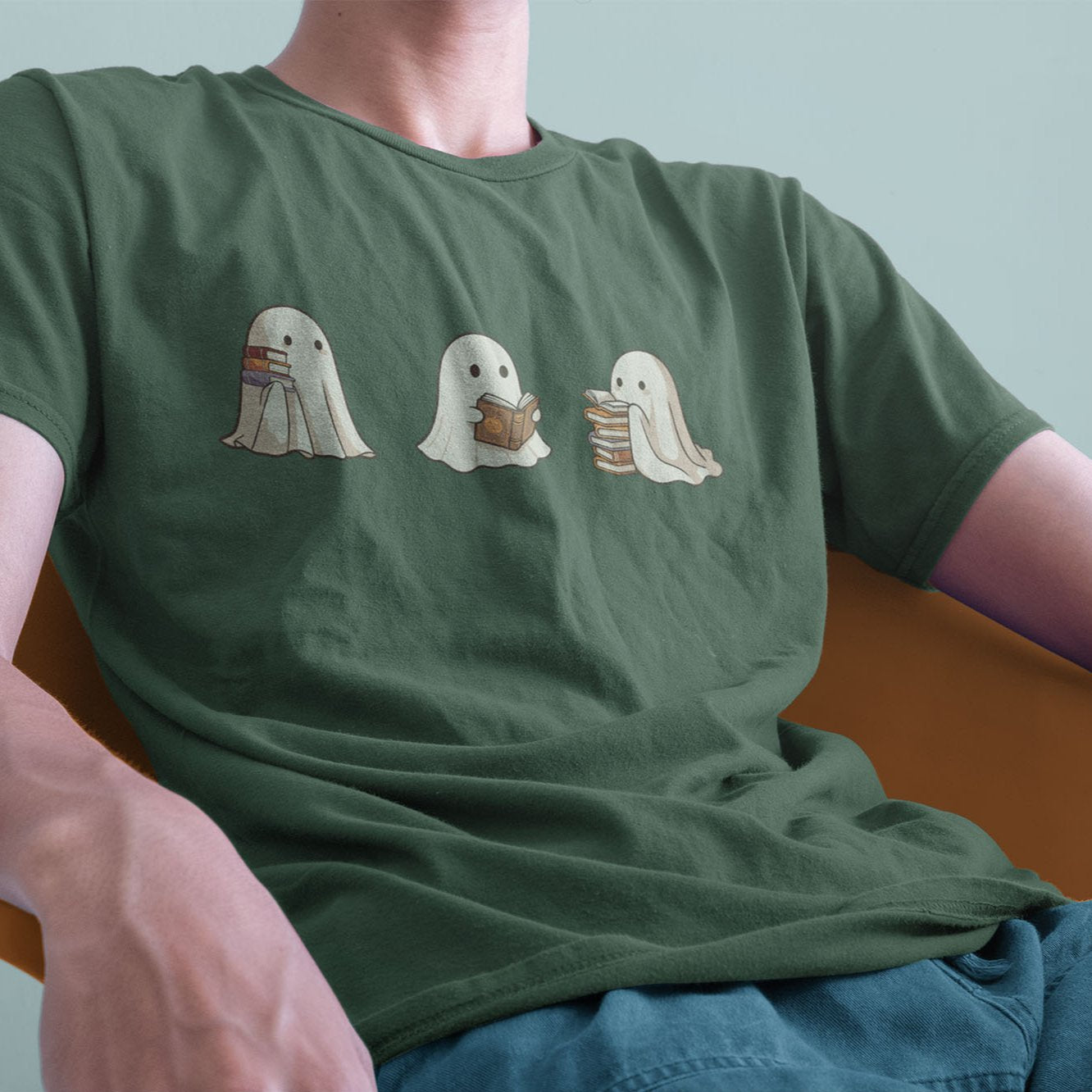 A male model leaning back on a chair, wearing a vintage Comfort Colors Tshirt in Moss Green color. The retro design shows three cute little bedsheet ghosts reading books together, in a fun cartoonish art style.