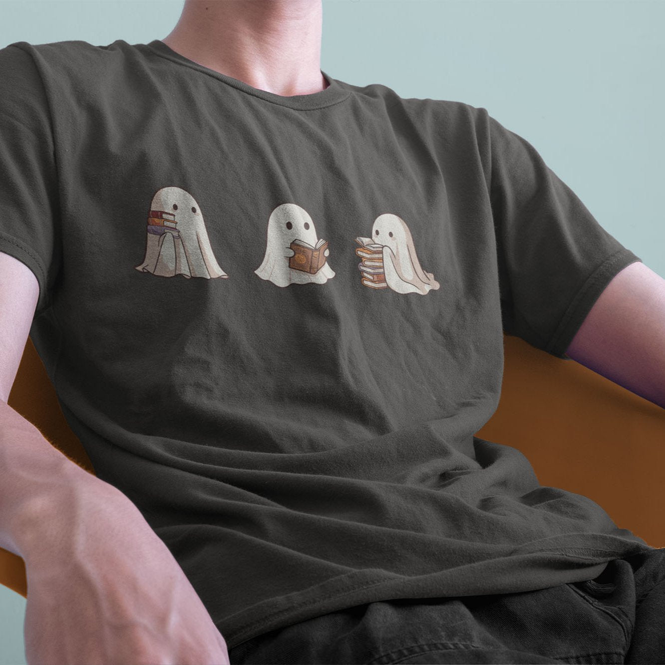 A male model leaning back on a chair, wearing a vintage Comfort Colors Tshirt in Pepper Gray color. The retro design shows three cute little bedsheet ghosts reading books together, in a fun cartoonish art style.