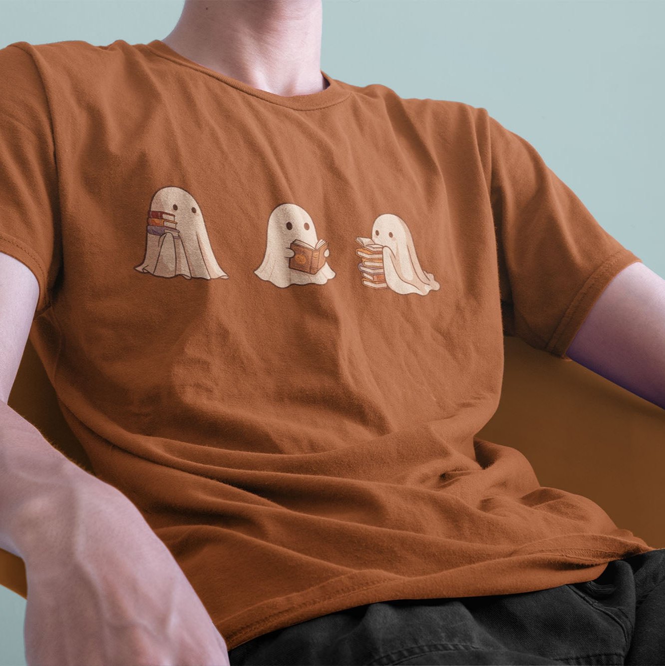 A male model leaning back on a chair, wearing a vintage Comfort Colors Tshirt in Yam Orange color. The retro design shows three cute little bedsheet ghosts reading books together, in a fun cartoonish art style.