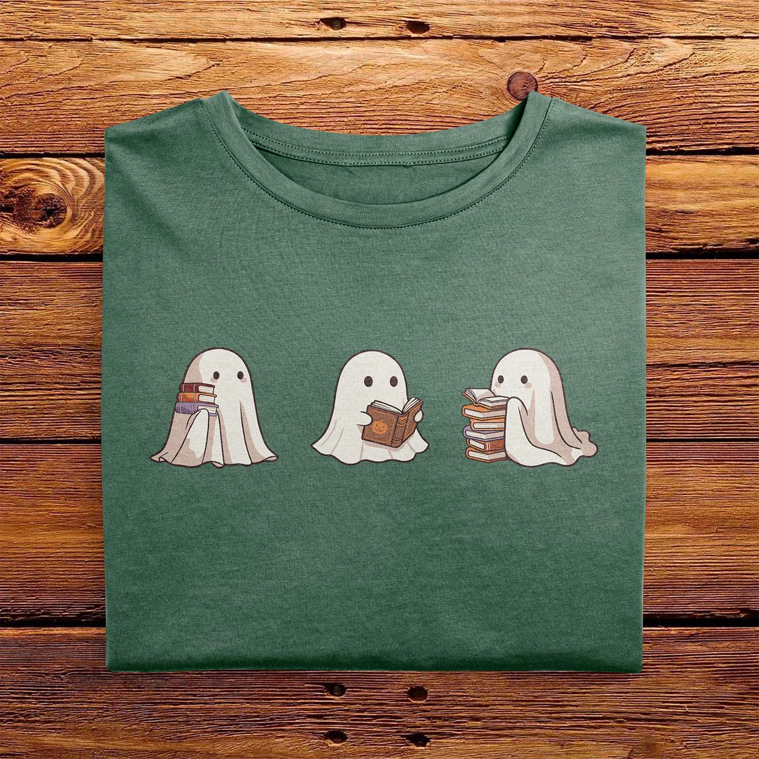 A nicely folded Comfort Colors shirt in Moss Green, laying on a wooden table. The retro design shows three cute little bedsheet ghosts reading books together, in a fun cartoonish art style, giving Fall vibes.