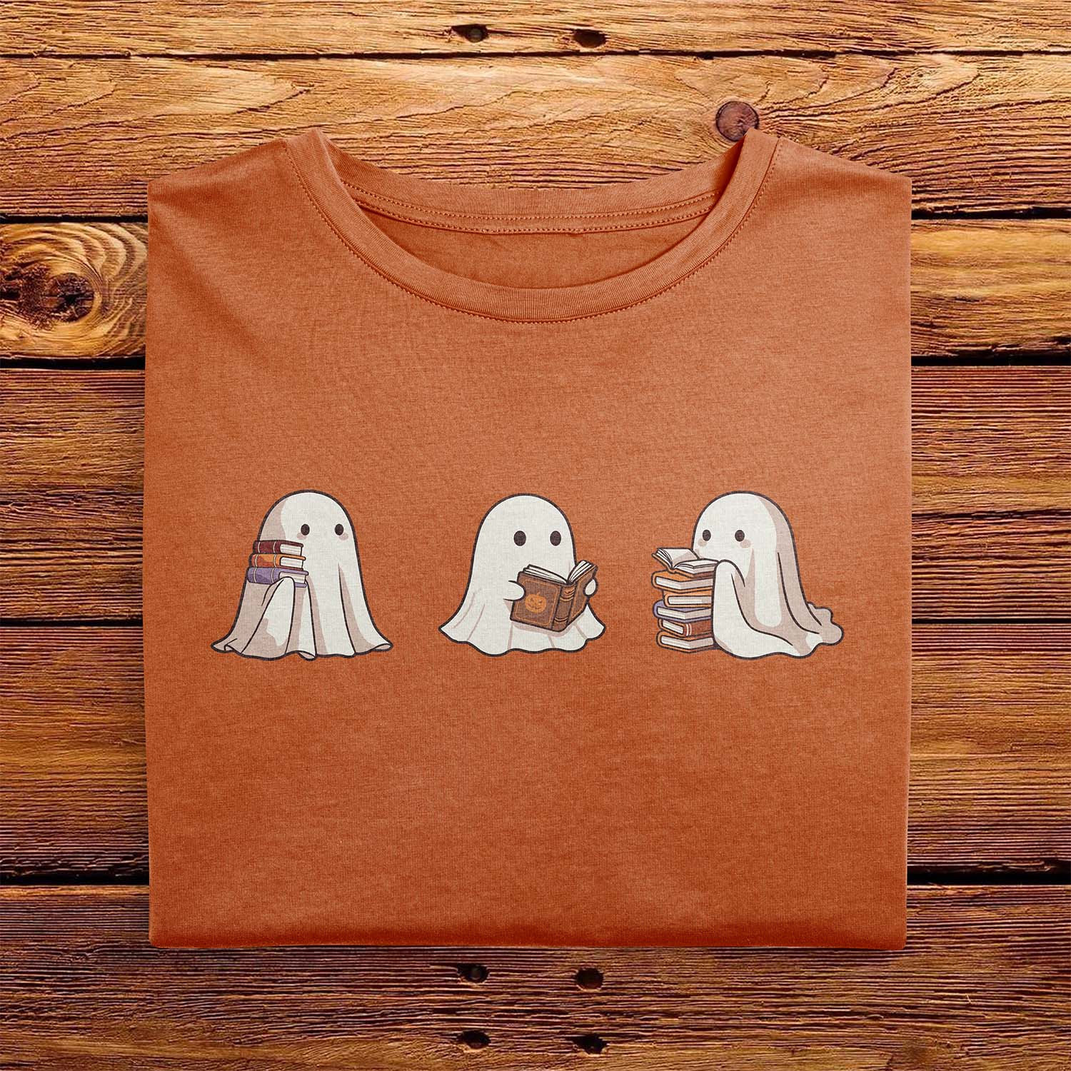 A nicely folded Comfort Colors shirt in Yam Orange, laying on a wooden table. The retro design shows three cute little bedsheet ghosts reading books together, in a fun cartoonish art style, giving Fall vibes.