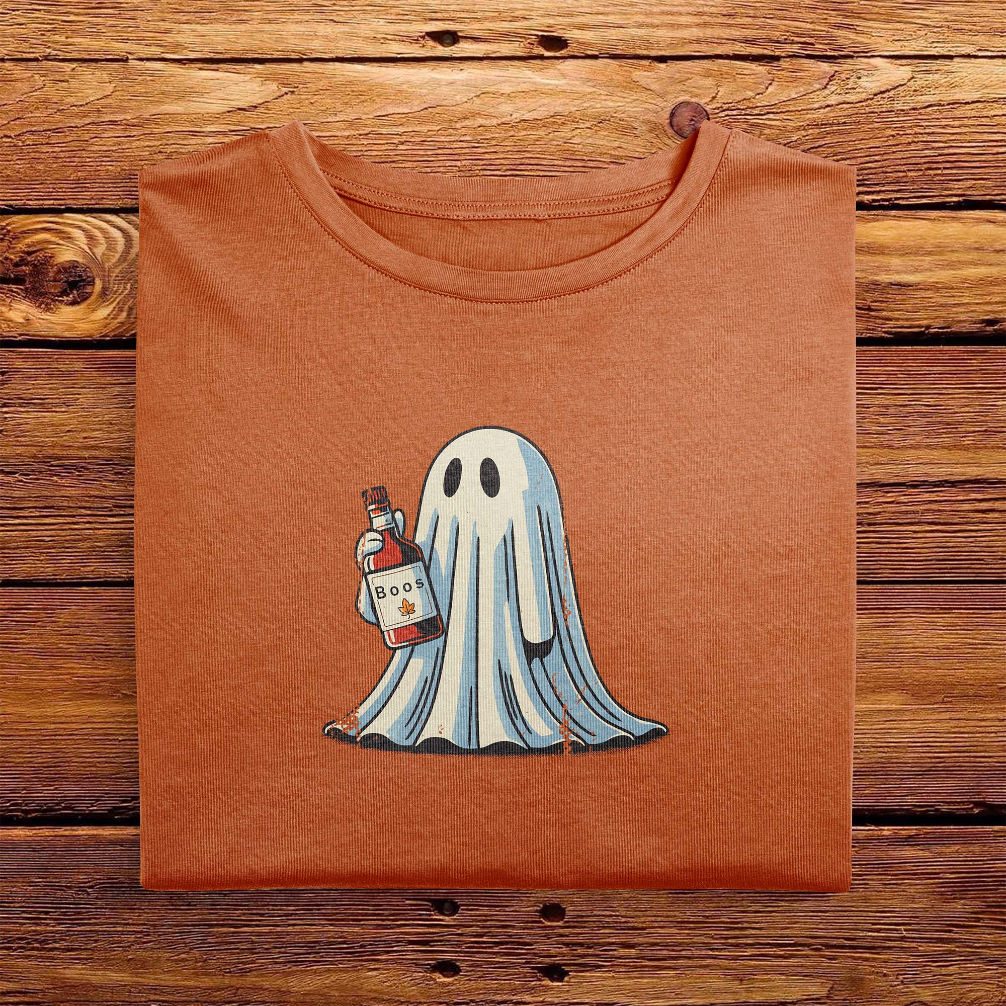 A folded vintage Comfort Colors Tshirt in Yam Orange color, laying on a wooden table. The retro design features a cute bedsheet ghost holding a bottle of liquor that says Boos.