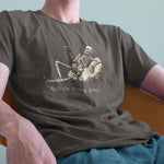 A closeup of a model in a chair wearing a comfort colors Tshirt in Pepper Gray color. The vintage includes a relaxed skeleton leaned back on a boulder, smoking a joint. There is text below it that says Built On Strong Joints.