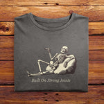 A folded Comfort Colors Vintage Tshirt in Pepper Gray color laying on a wooden table. The design includes a relaxed skeleton leaned back on a boulder, smoking a joint. There is text below it that says Built On Strong Joints.