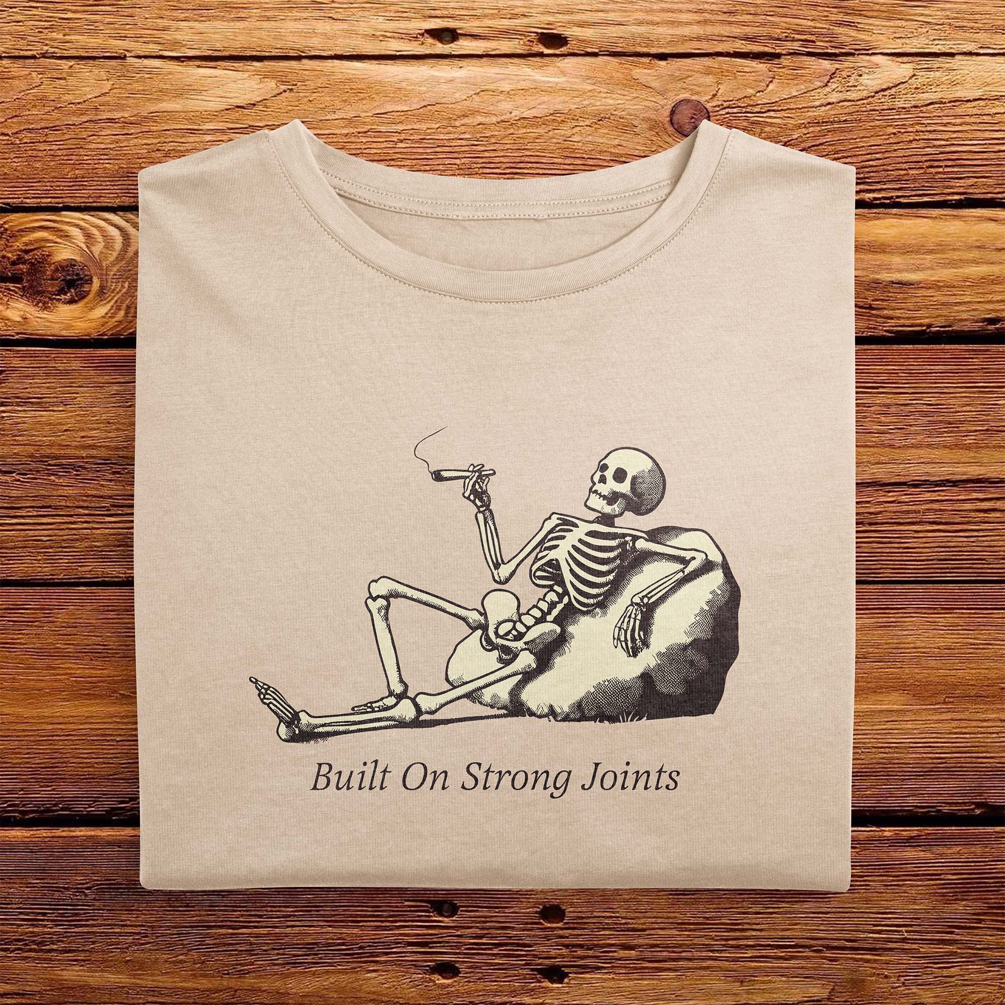 A folded Comfort Colors Vintage Tshirt in Ivory color laying on a wooden table. The design includes a relaxed skeleton leaned back on a boulder, smoking a joint. There is text below it that says Built On Strong Joints.