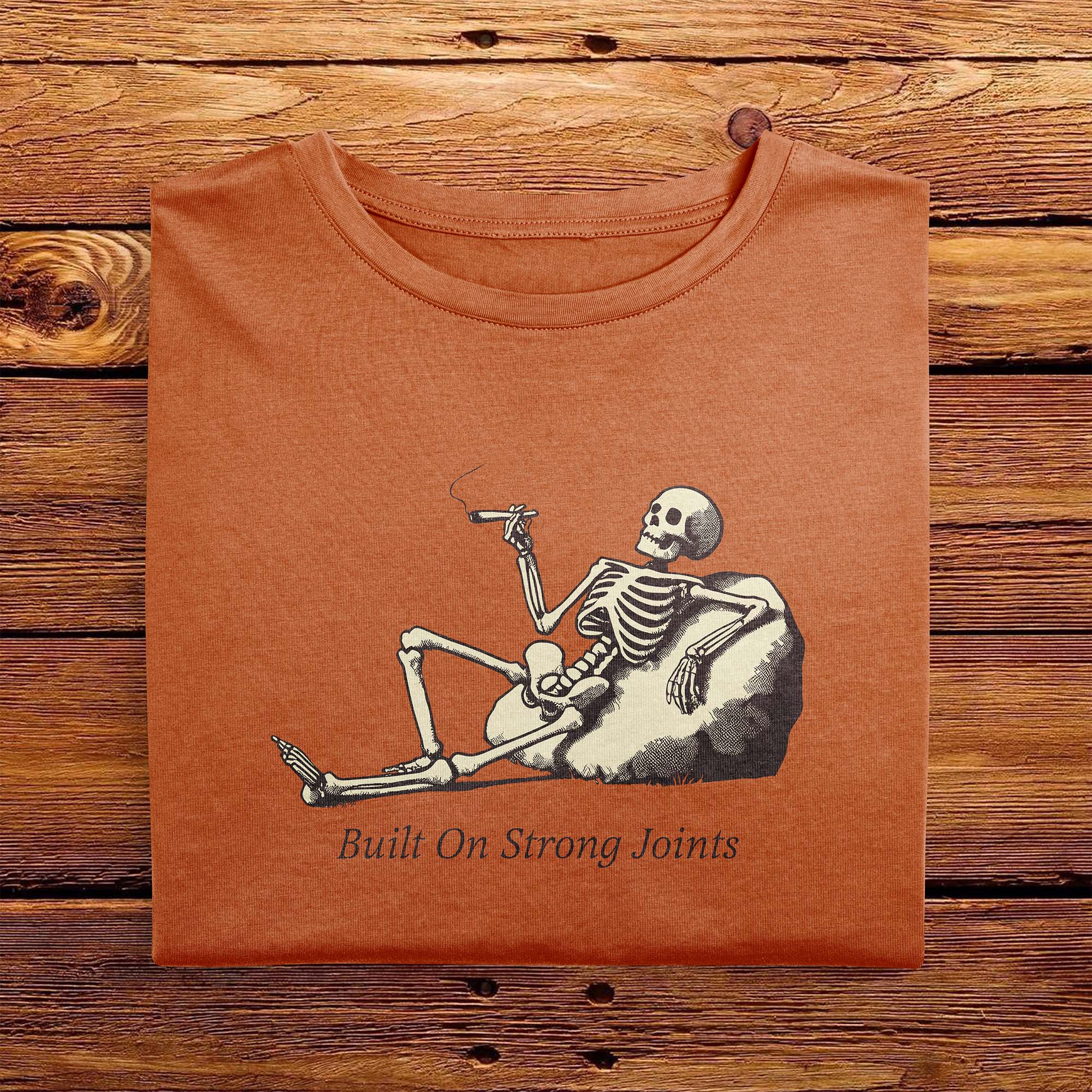 A folded Comfort Colors Vintage Tshirt in Yam Orange color laying on a wooden table. The design includes a relaxed skeleton leaned back on a boulder, smoking a joint. There is text below it that says Built On Strong Joints.