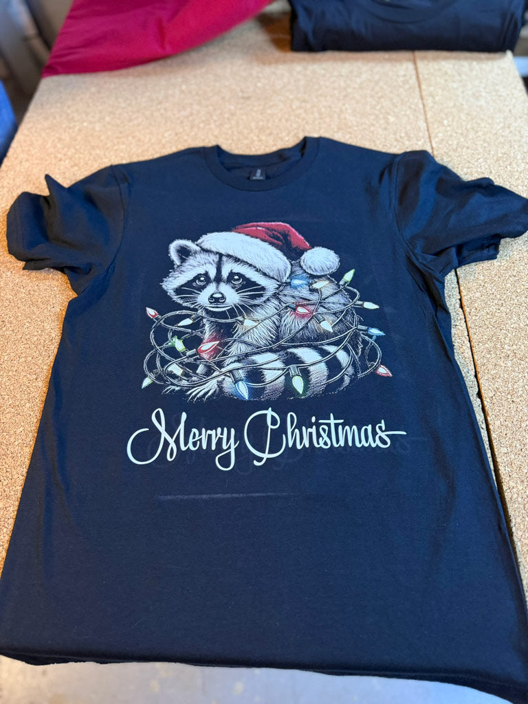 Black t-shirt printed with a detailed raccoon wearing a Santa hat tangled in colorful Christmas lights above scripted “Merry Christmas” text.