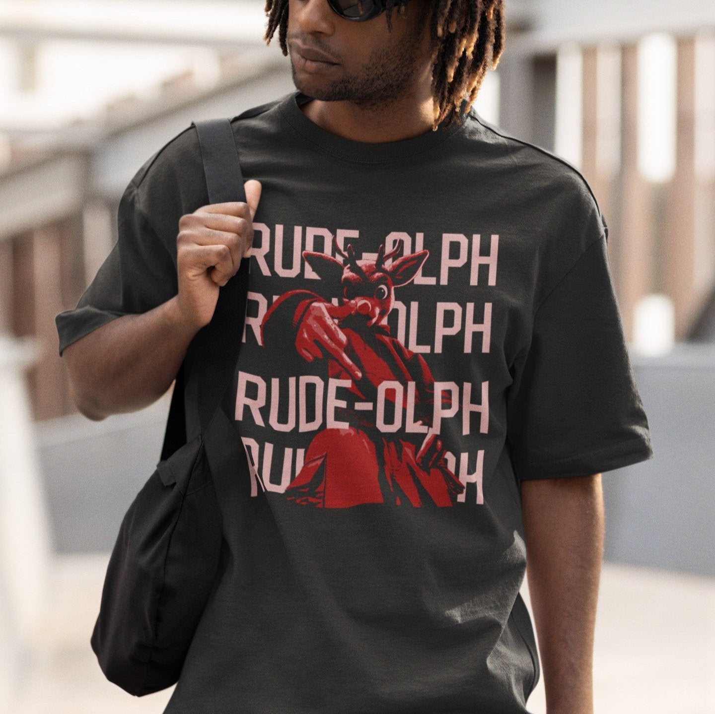 A model wearing a vintage baggy grey comfort colors shirt. The retro design features Rudolph the Red Nosed Reindeer in a y2k merch artstyle, posed in a rude manner. There is text that says Rude-olph, a funny pun with his name to go with the graphic