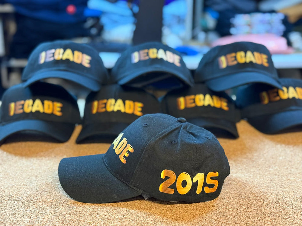 Black baseball caps stacked on a table, each embroidered “DECADE” in metallic gold, with one cap in front showing “2015” in gold on its side panel.