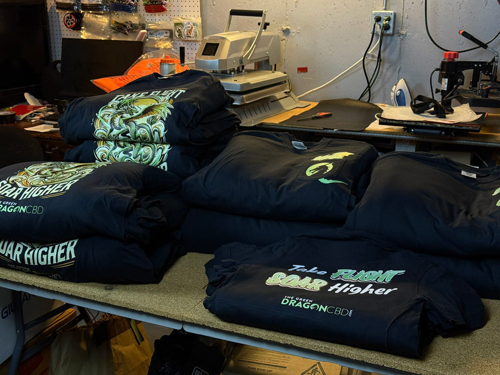 Stacks of black custom t-shirts on a workshop table featuring neon green dragon artwork and “Take Flight Soar Higher” text with The Green Dragon CBD branding under a heat-press machine.