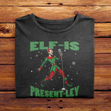 A Black Folded Comfort Colors Tshirt featuring a vintage-style illustration of an elf singing into a microphone. The green text above and below the elf reads ELF-IS PRESENT-LEY, in reference to Elvis Presley, and cute festive snowflakes scattered around.