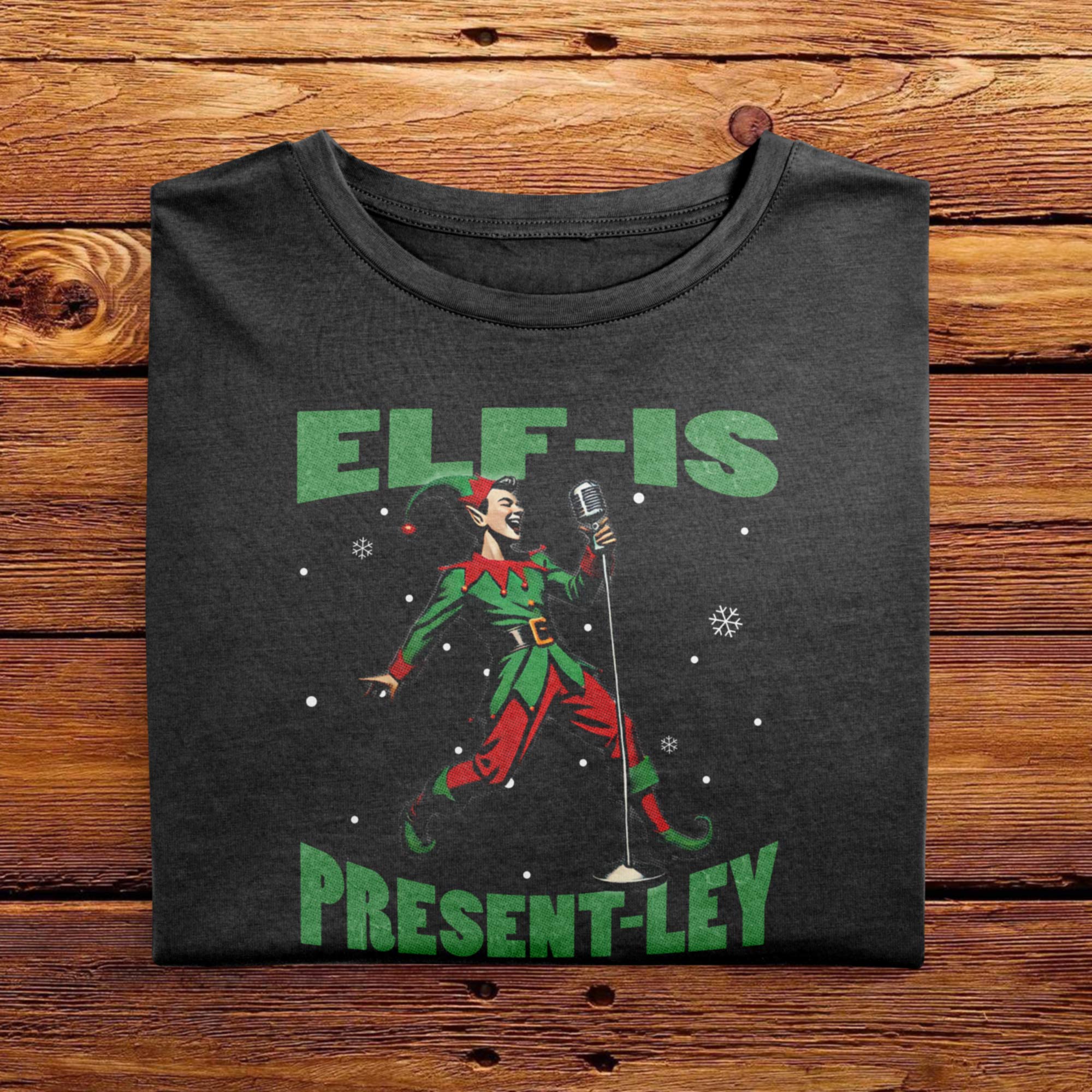 A Black Folded Comfort Colors Tshirt featuring a vintage-style illustration of an elf singing into a microphone. The green text above and below the elf reads ELF-IS PRESENT-LEY, in reference to Elvis Presley, and cute festive snowflakes scattered around.