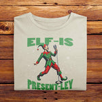 A Ivory White Folded Comfort Colors Tshirt featuring a vintage-style illustration of an elf singing into a microphone. The green text above and below the elf reads ELF-IS PRESENT-LEY, in reference to Elvis Presley, and cute festive snowflakes scattered around.