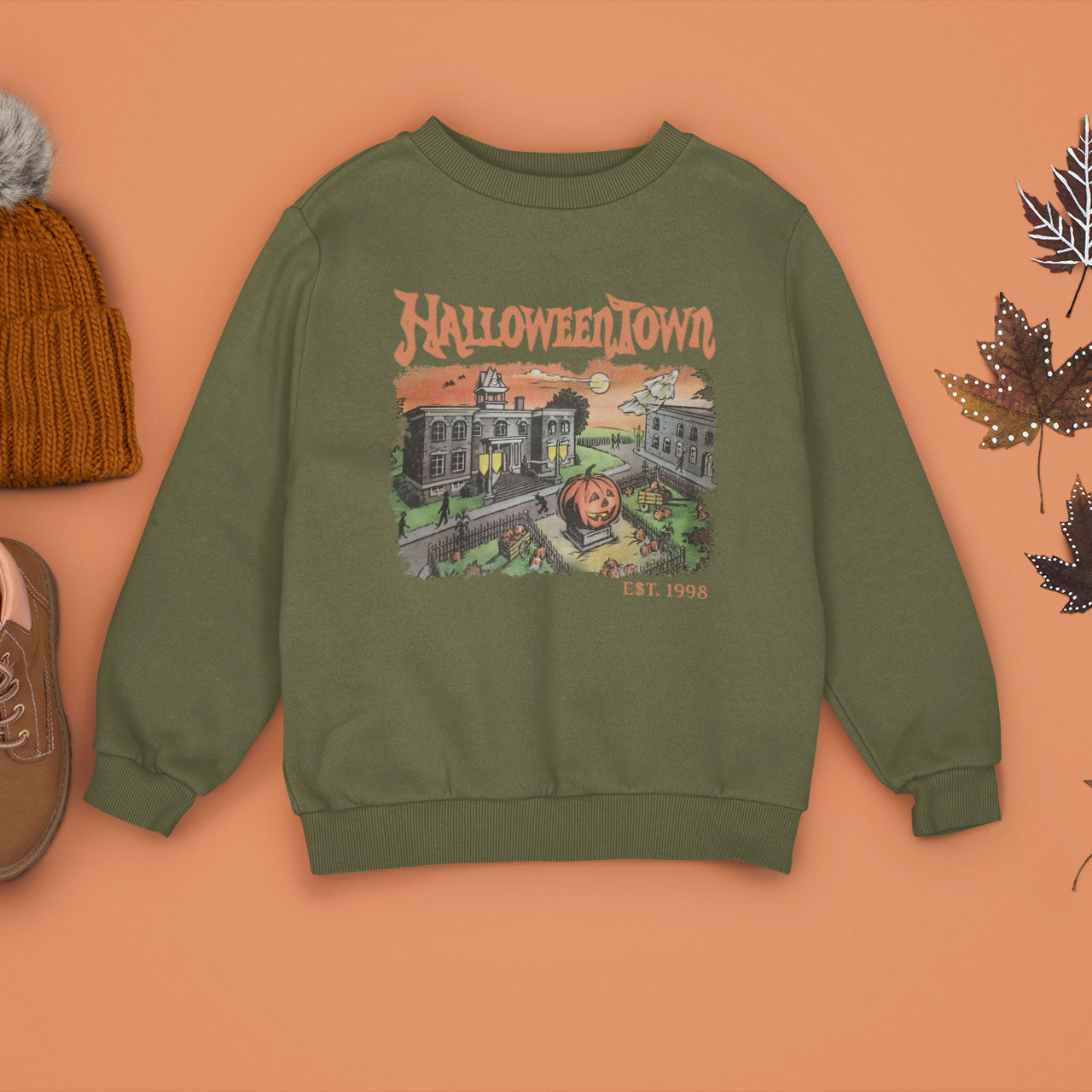 A vintage green sweatshirt with a retro graphic print of a spooky town with a large pumpkin in front of a building. The text on the sweatshirt reads Halloweentown Est. 1998