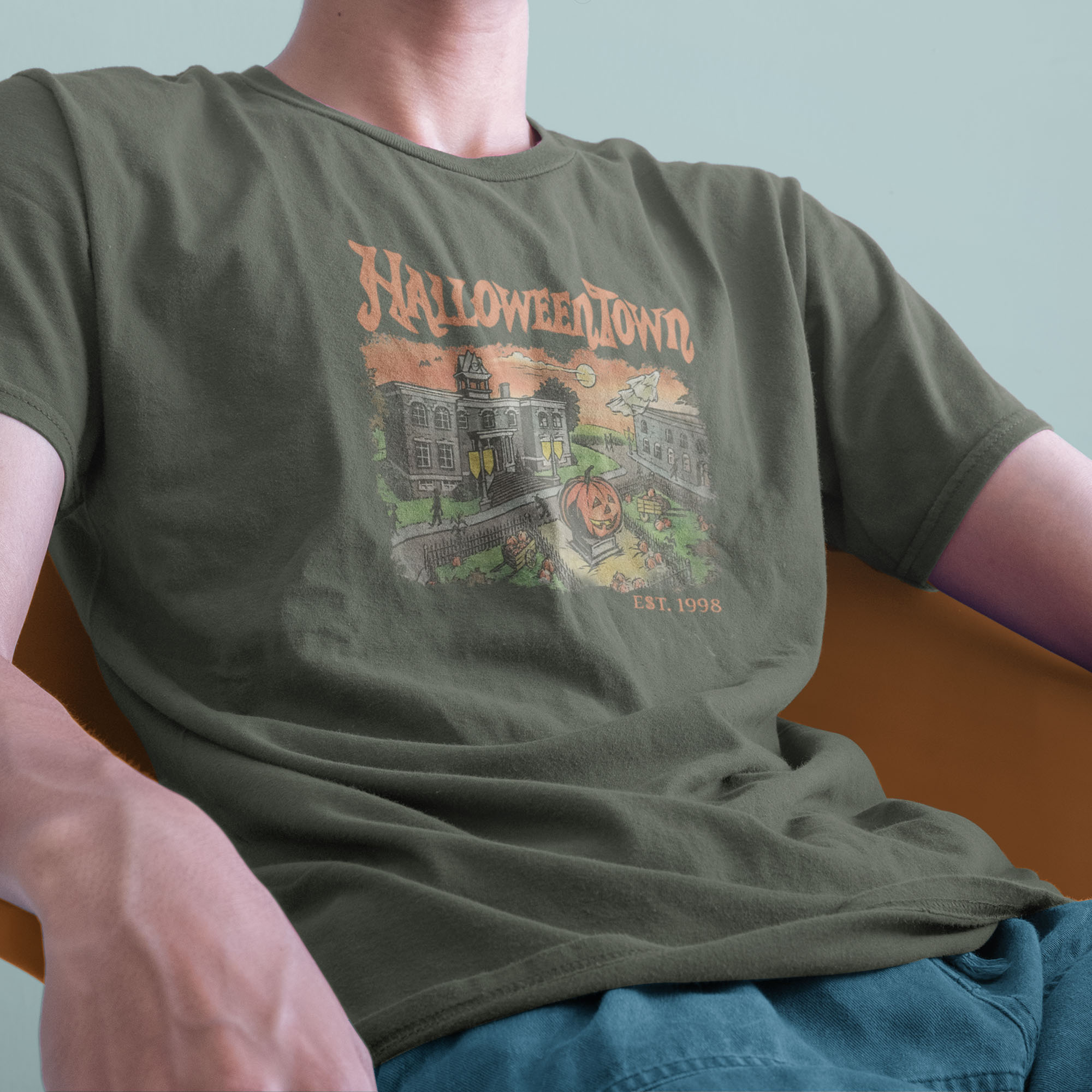 Amale model sitting down wearing a vintage Comfort Colors Tshirt in Moss Green with a retro graphic print of a spooky town with a large pumpkin in front of a building. The text on the sweatshirt reads Halloweentown Est. 1998