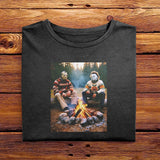 A Black folded Comfort Colors Tshirt, laying on a wooden table. The vintage design features Jason Vorhees from Friday the 13th, and Michael Meyers from Halloween, both in cozy sweaters sitting around a campfire roasting marshmallows.