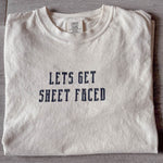 A Comfort Colors Tshirt in Ivory White color folded nicely with a modern wooden gray backdrop. There is a funny halloween themed pun on the front of the shirt that says Lets Get Sheet Faced.