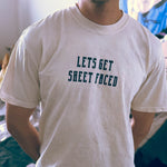 A male model wearing a Comfort Colors Tshirt in Ivory White color with his hands behind his back. There is a funny halloween themed pun on the front of the shirt that says Lets Get Sheet Faced.