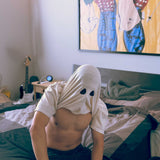 A male model wearing a Comfort Colors Tshirt in Ivory White sitting down on a bed. The shirt is pulled up above his head, revealing two black spots resembling the eyes of a bedsheet ghost. This shirt has a fun halloween themed design.