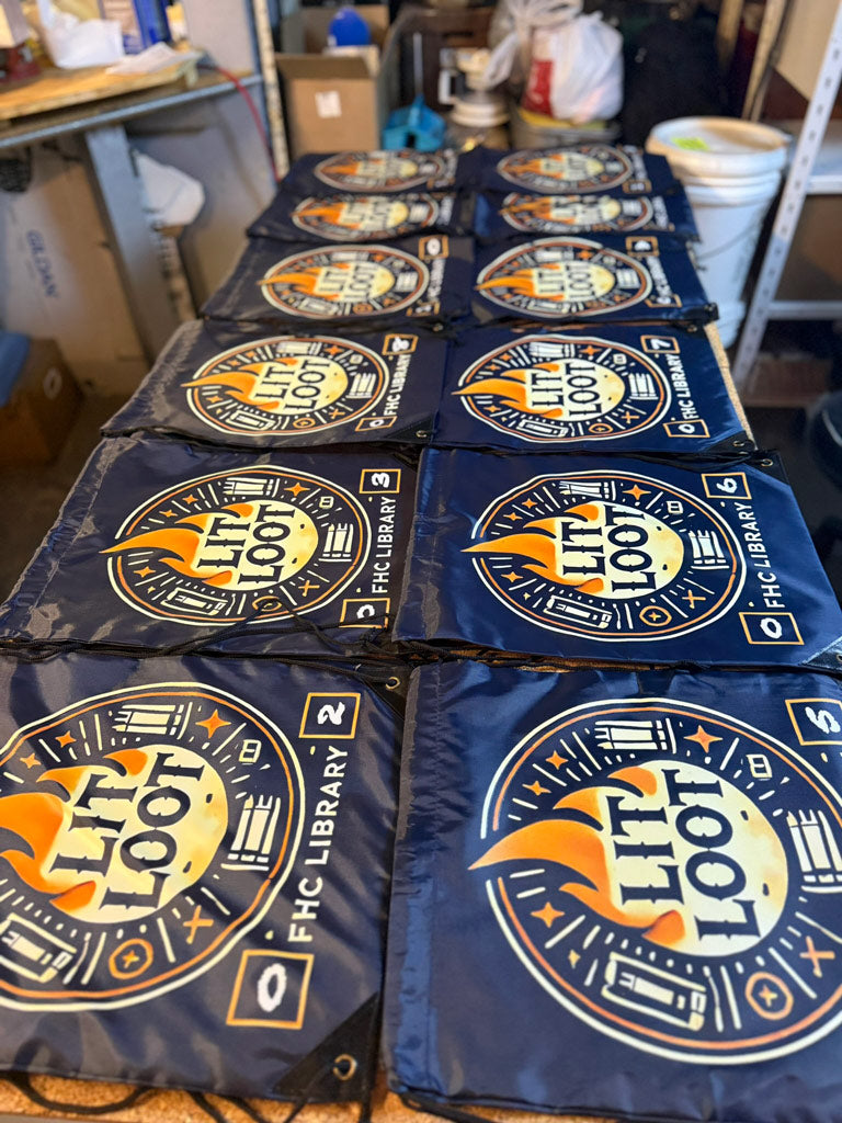 Navy drawstring backpacks numbered 1–8 laid out on a worktable, each featuring a circular “LIT LOOT” flame-and-book logo and FHC Library branding.