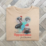 An off white folded tshirt with a festive design featuring Moo Deng, a baby pygmy hippo wearing Santa hats in three poses with a full moon behind. The text reads I want a Hippopotamus for Christmas, blending holiday fun with festive cheer.