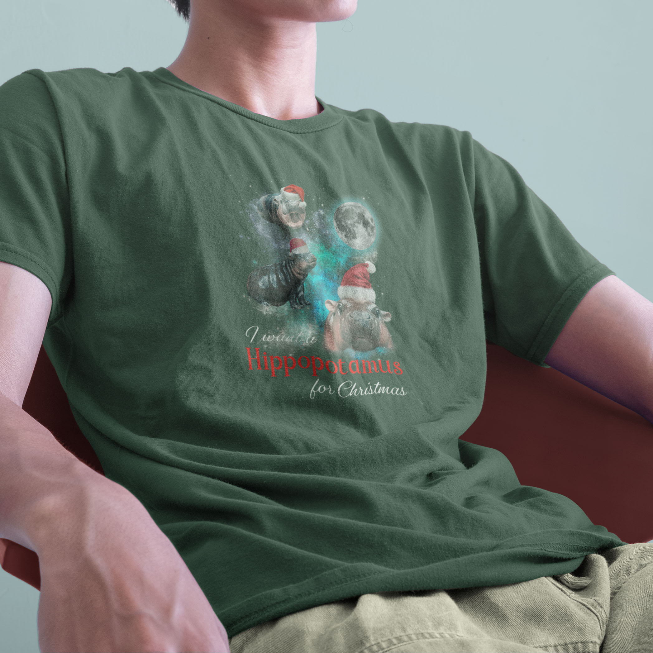 A model wearing a green tshirt with a festive design featuring Moo Deng, a baby pygmy hippo wearing Santa hats in three poses with a full moon behind. The text reads I want a Hippopotamus for Christmas, blending holiday fun with festive cheer.