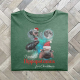 A green folded tshirt with a festive design featuring Moo Deng, a baby pygmy hippo wearing Santa hats in three poses with a full moon behind. The text reads I want a Hippopotamus for Christmas, blending holiday fun with festive cheer.