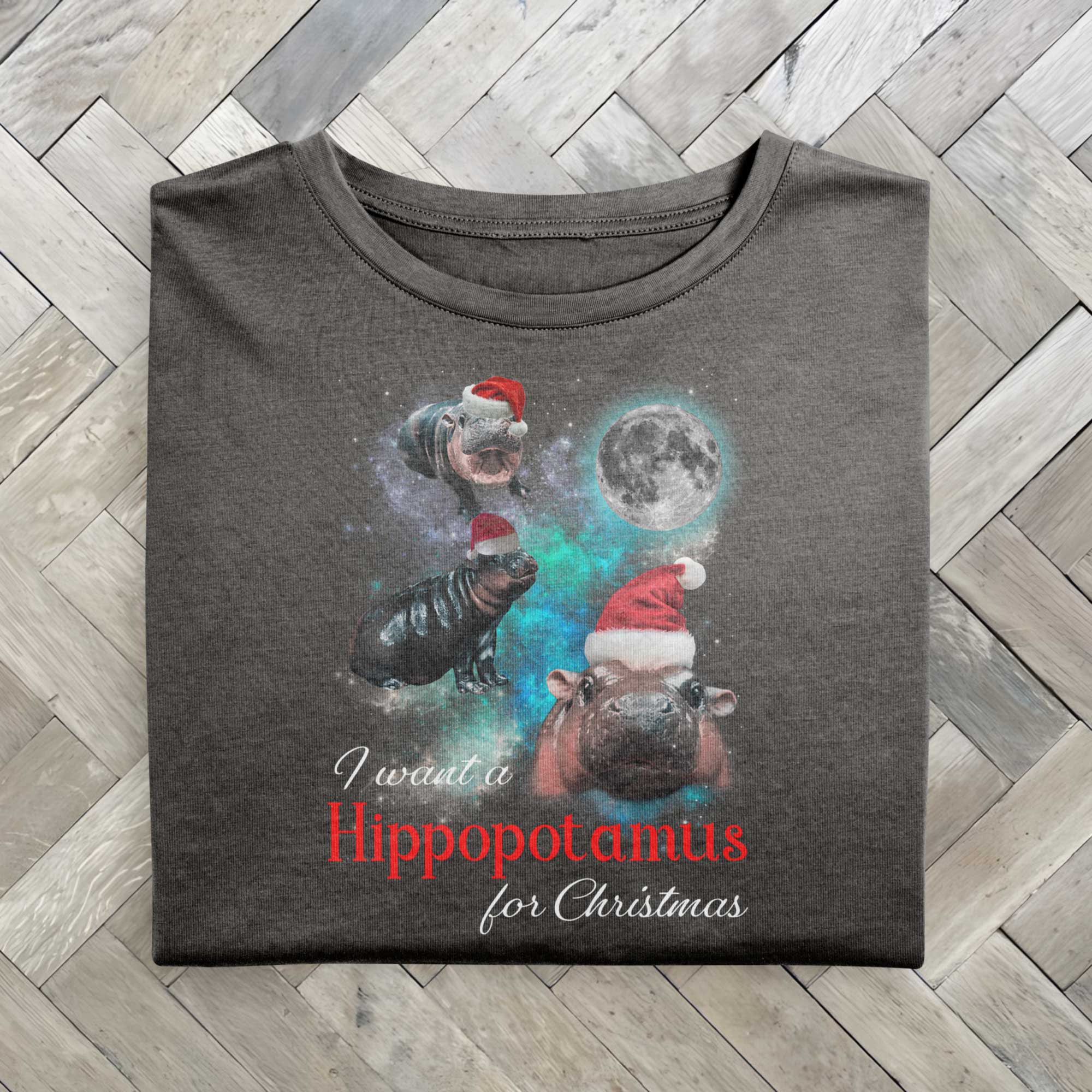 A gray folded tshirt with a festive design featuring Moo Deng, a baby pygmy hippo wearing Santa hats in three poses with a full moon behind. The text reads I want a Hippopotamus for Christmas, blending holiday fun with festive cheer.