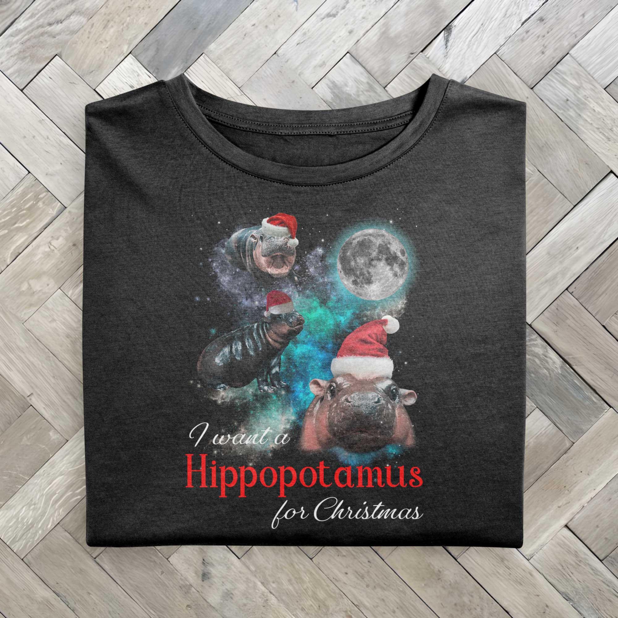 A black folded tshirt with a festive design featuring Moo Deng, a baby pygmy hippo wearing Santa hats in three poses with a full moon behind. The text reads I want a Hippopotamus for Christmas, blending holiday fun with festive cheer.