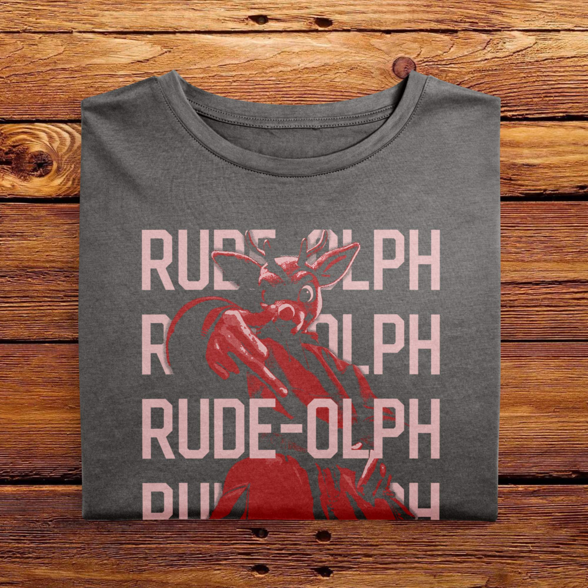 A vintage folded Pepper Grey comfort colors shirt. The festive design features Rudolph the Red Nosed Reindeer in a y2k merch artstyle, posed in a rude manner. There is text that says Rude-olph, a funny festive pun with his name to go with the graphic