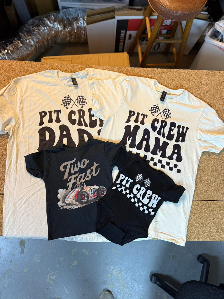 Cream-colored “Pit Crew Daddy” and “Pit Crew Mama” tees with checkered flag graphics, alongside a black “Two Fast” toddler tee and matching “Pit Crew” baby onesie.