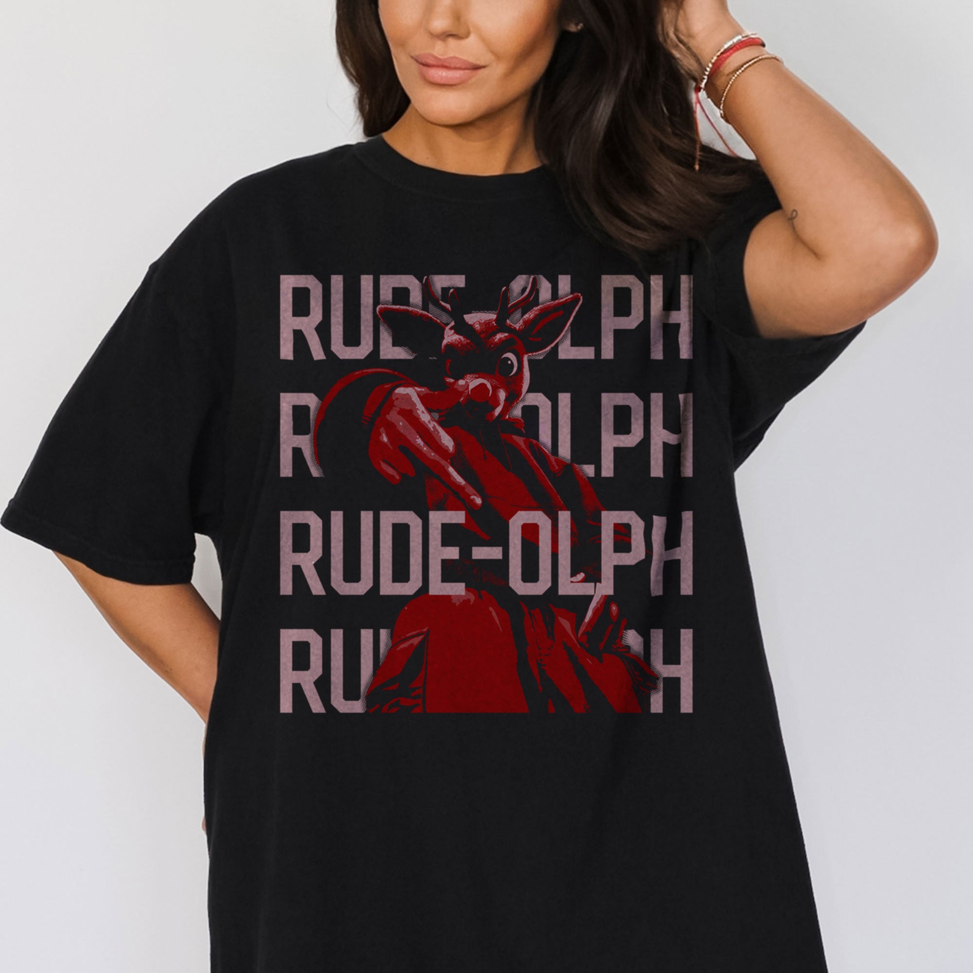 A model wearing a vintage baggy black comfort colors shirt. The retro design features Rudolph the Red Nosed Reindeer in a y2k merch artstyle, posed in a rude manner. There is text that says Rude-olph, a funny pun with his name to go with the graphic