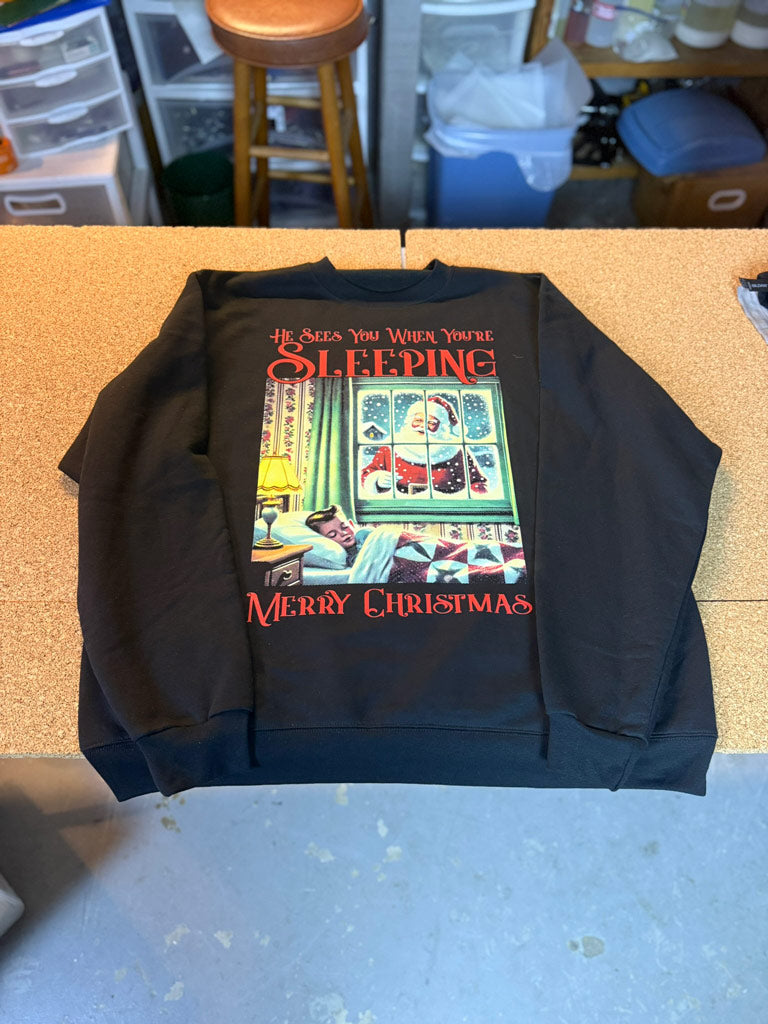 Black crewneck sweatshirt printed with vintage Santa peering in a child’s bedroom window above “He Sees You When You’re Sleeping” and “Merry Christmas” text in red.