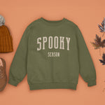 A Fresh Olive Green Crewneck Champion Power blend Sweatshirt, on an earthy autumn backdrop. This design features the words: Spooky Season, in an off white scholarly university font.