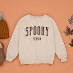 A sand white Crewneck Champion Power blend Sweatshirt, on an earthy autumn backdrop. This design features the words: Spooky Season, in an off white scholarly university font.