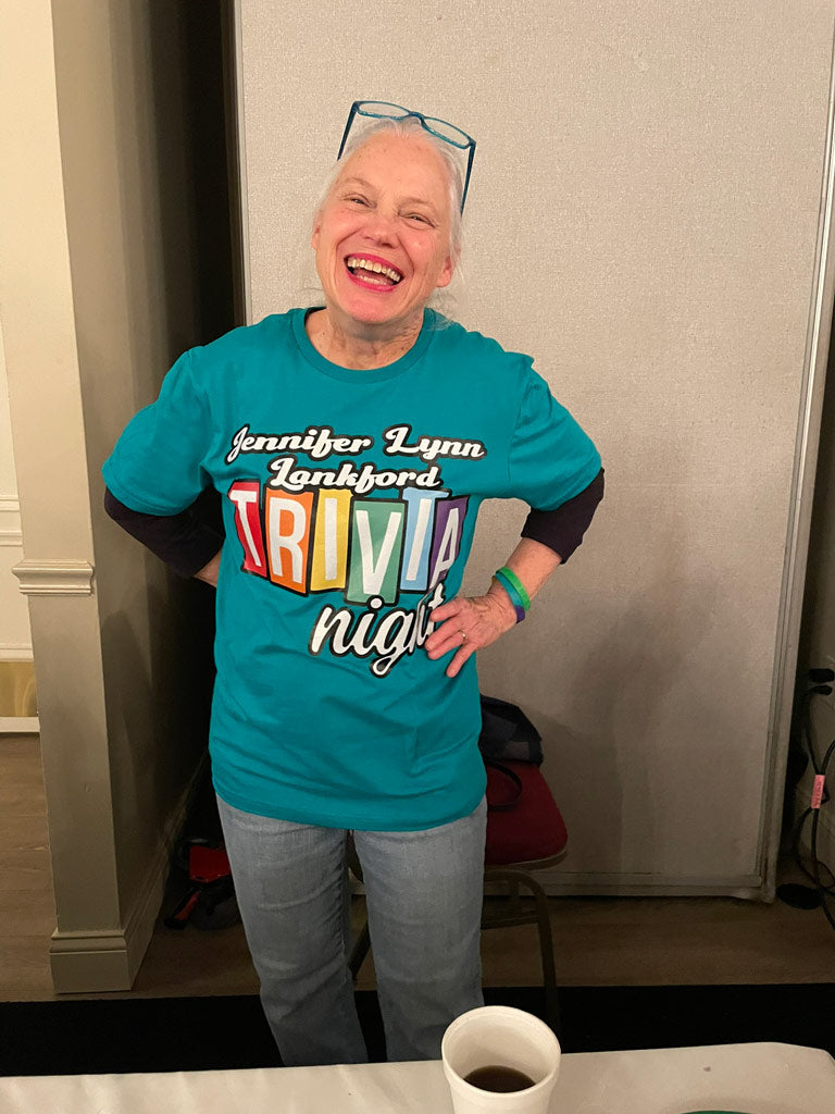 Smiling woman wearing a teal “Jennifer Lynn Lankford Trivia Night” tee in rainbow letters, standing at a community event table.