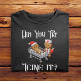 A Black Comfort Colors Tshirt featuring a playful design of a gingerbread nurse and injured gingerbread man in a hospital bed. The text reads Did You Try Icing It? Perfect holiday gift for nurses, bakers, or those with a sense of humor.