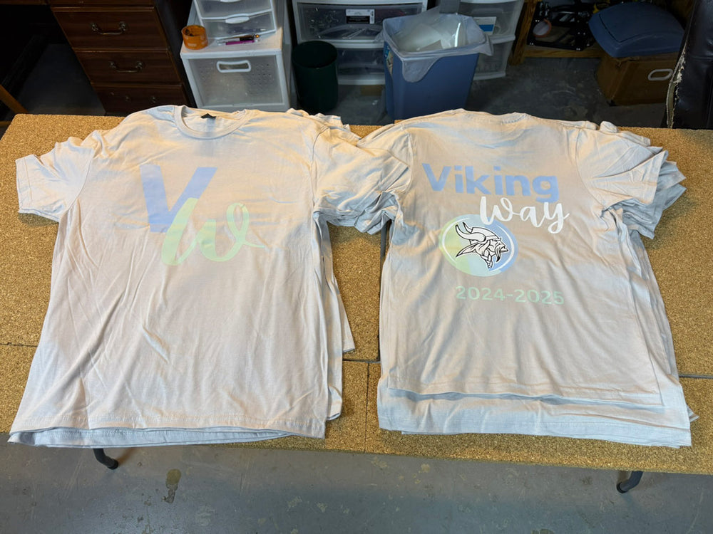 Light gray tees laid out front and back: front shows pastel “Vw” monogram; back reads “Viking Way 2024–2025” above a Viking helmet emblem.