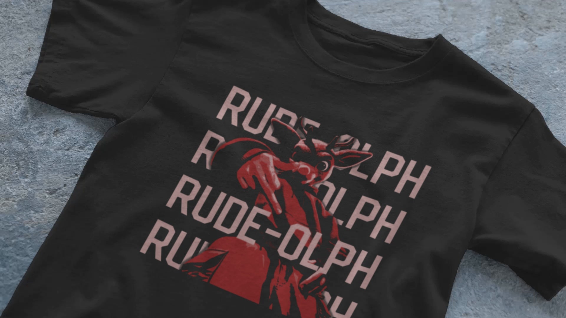 A video of a laid out black comfort colors shirt. The retro design features Rudolph the Red Nosed Reindeer in a y2k merch artstyle, posed in a rude manner. There is text that says Rude-olph, a funny festive pun with his name to go with the graphic